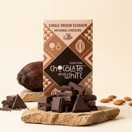A boxed bar of chocolate stands upright on a stone slab with several chocolate pieces placed in front. A cocoa pod and a few almonds are visible in the background on a warm cream studio backdrop.